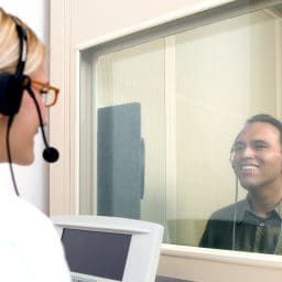 Man receiving a hearing test in a hearing booth looking through a glass window to a female audiologist